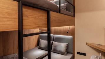 Una cama individual con estructura de madera y ropa de cama gris, una mesita de noche con lámpara, y una maleta en un estante.