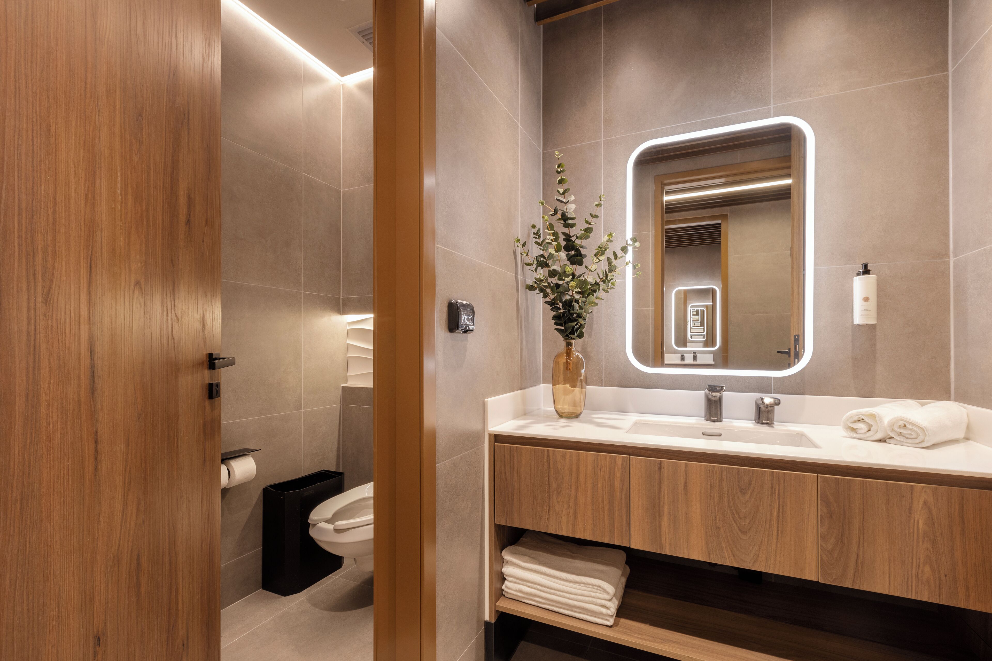 A modern bathroom with a wooden vanity, a large mirror, and a vase with flowers.