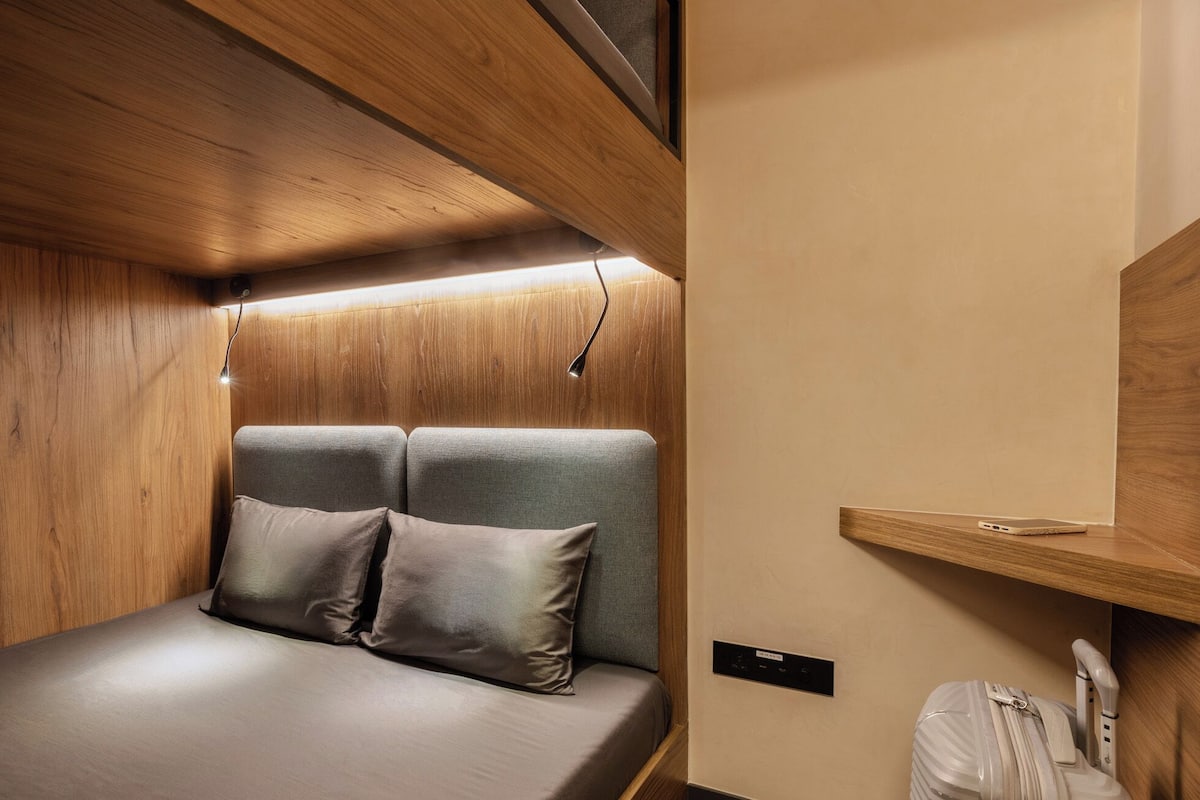 Ein gemütliches, gut beleuchtetes Zimmer mit einem Bett, zwei Kissen, einer kleinen Ablagefläche und einem Koffer.