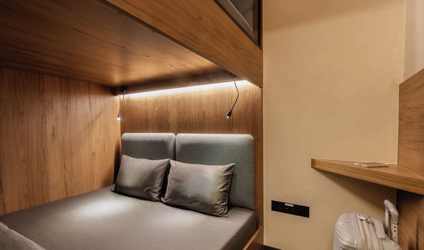 A compact, well-lit cabin with a bed, two pillows, a small shelf, and a suitcase. - Sleepover - Lima Airport, the only in-terminal sleep pods (Callao)