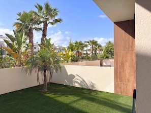 Un patio con palmeras, césped artificial, un muro blanco y una valla de madera.