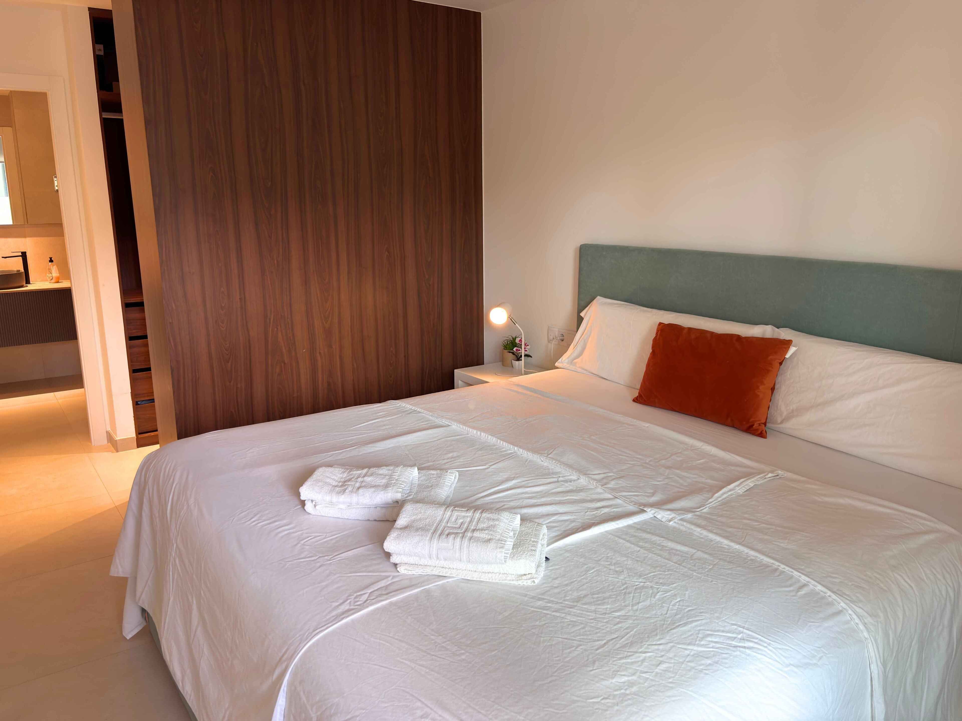A neatly made bed with white linens and a single orange pillow, a bedside lamp, and a wooden panel wall.