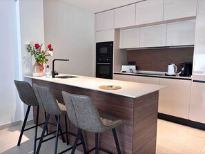 A modern kitchen with a central island, bar stools, a built-in oven, and a coffee maker.
