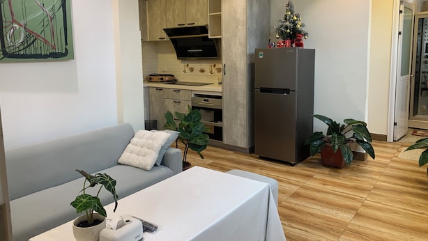 A modern living room with a sofa, a table with a tablecloth, and a refrigerator. There is a kitchen area with cabinets and a microwave. - Holly Apartment - Truc Bach Lake (Hanoi)