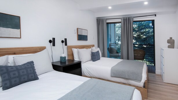A modern hotel room with two beds, a nightstand, a desk, and a view of the outdoors.