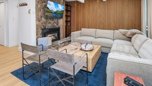 A modern living room with a stone fireplace, a large flat-screen TV, a wooden panel wall, a beige sectional sofa, a wooden coffee table, and a blue rug.