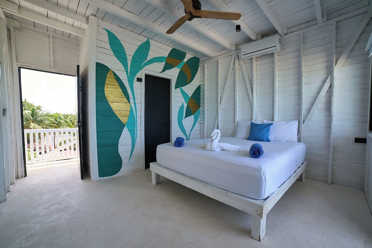 A bedroom with a bed, a ceiling fan, and a view of palm trees through an open door.