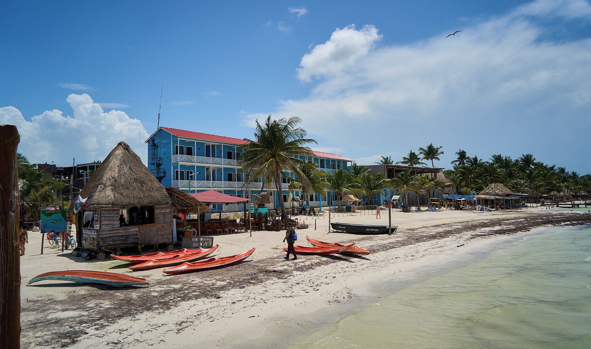 A beach with thatched-roof huts, a blue building with balconies, and several boats on the sand.