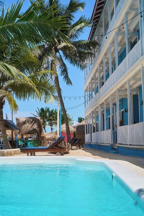 A pool area with lounge chairs, a thatched-roof hut, and a multi-story building with balconies.