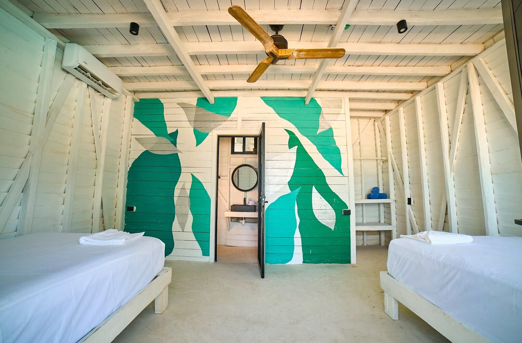 A room with two beds, a ceiling fan, a door leading to a bathroom, and a green abstract wall design.