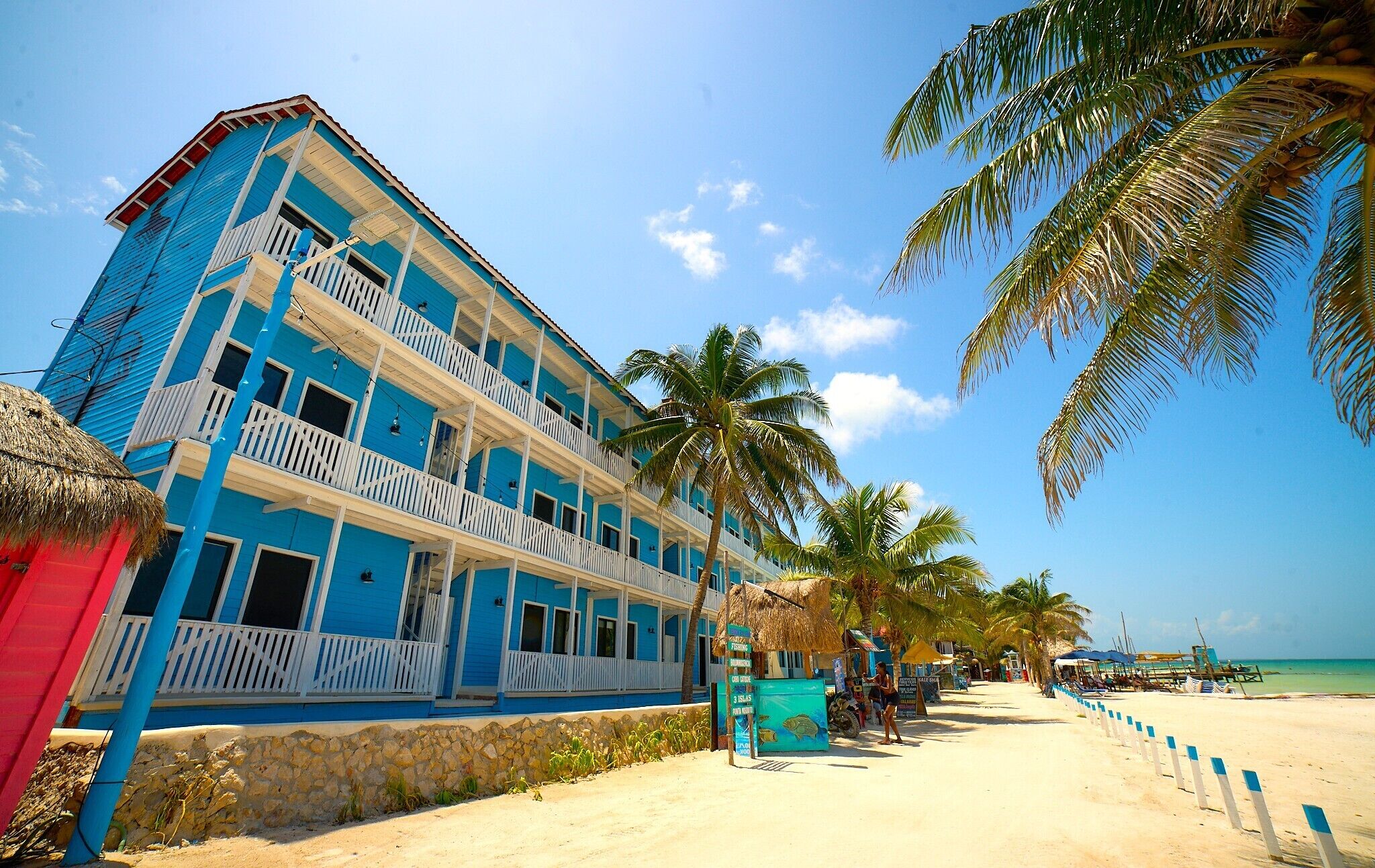 A multi-story building with balconies, a beach in the background, and palm trees.
