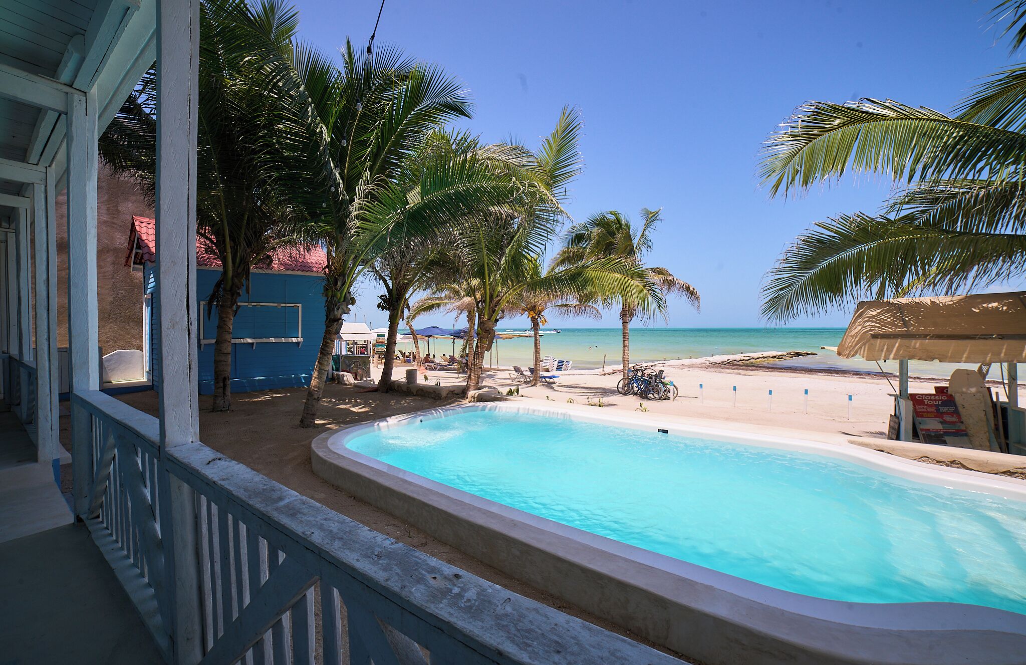 Vista de la piscina con cielo azul despejado, palmeras y una playa de arena.