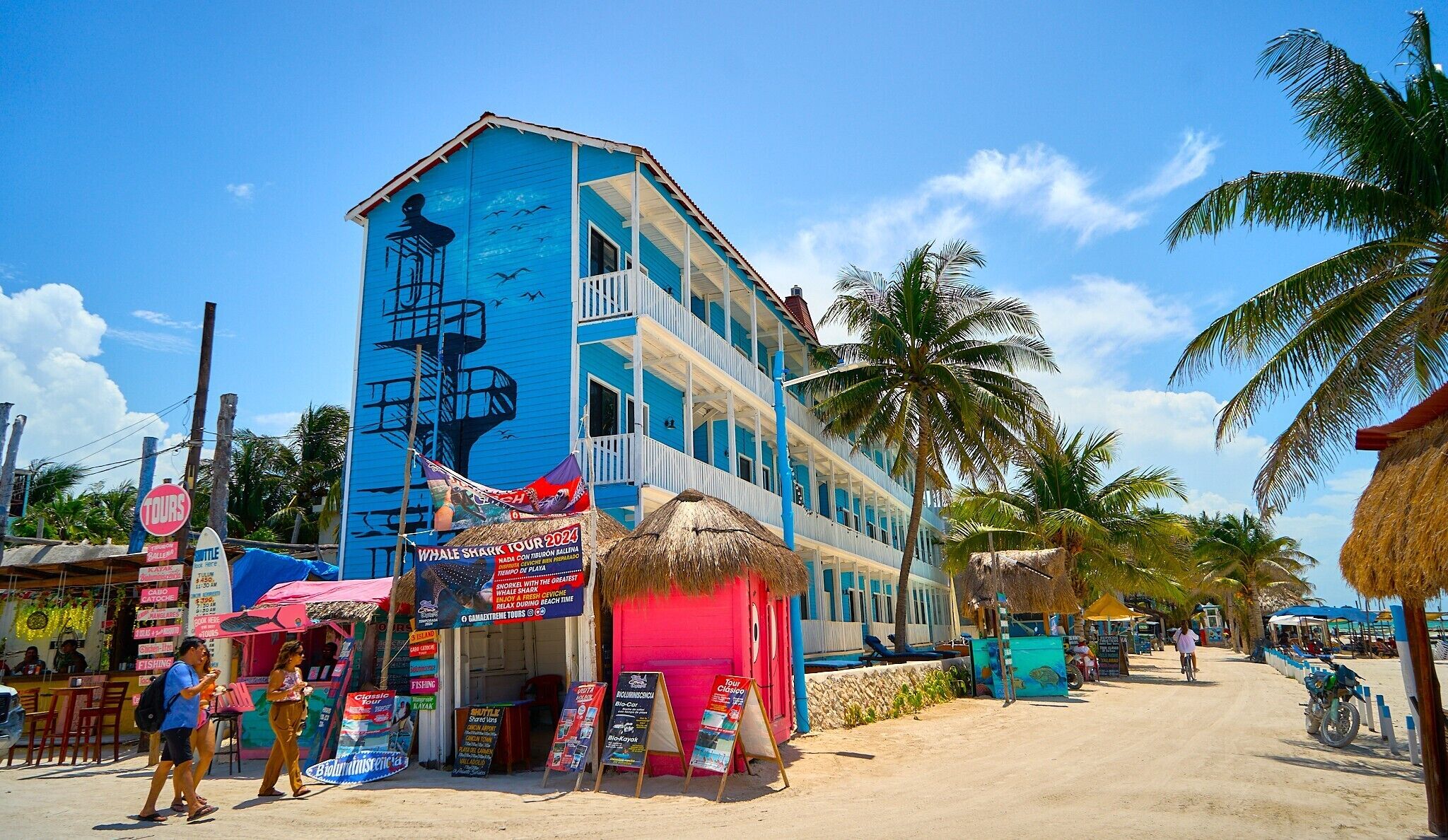 A multi-story building with balconies, a sign for whale shark tours, and a beachside location with palm trees and beachside activities.