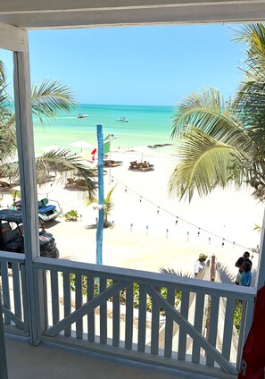 A beach view from a balcony with a railing, palm trees, and a clear sky.