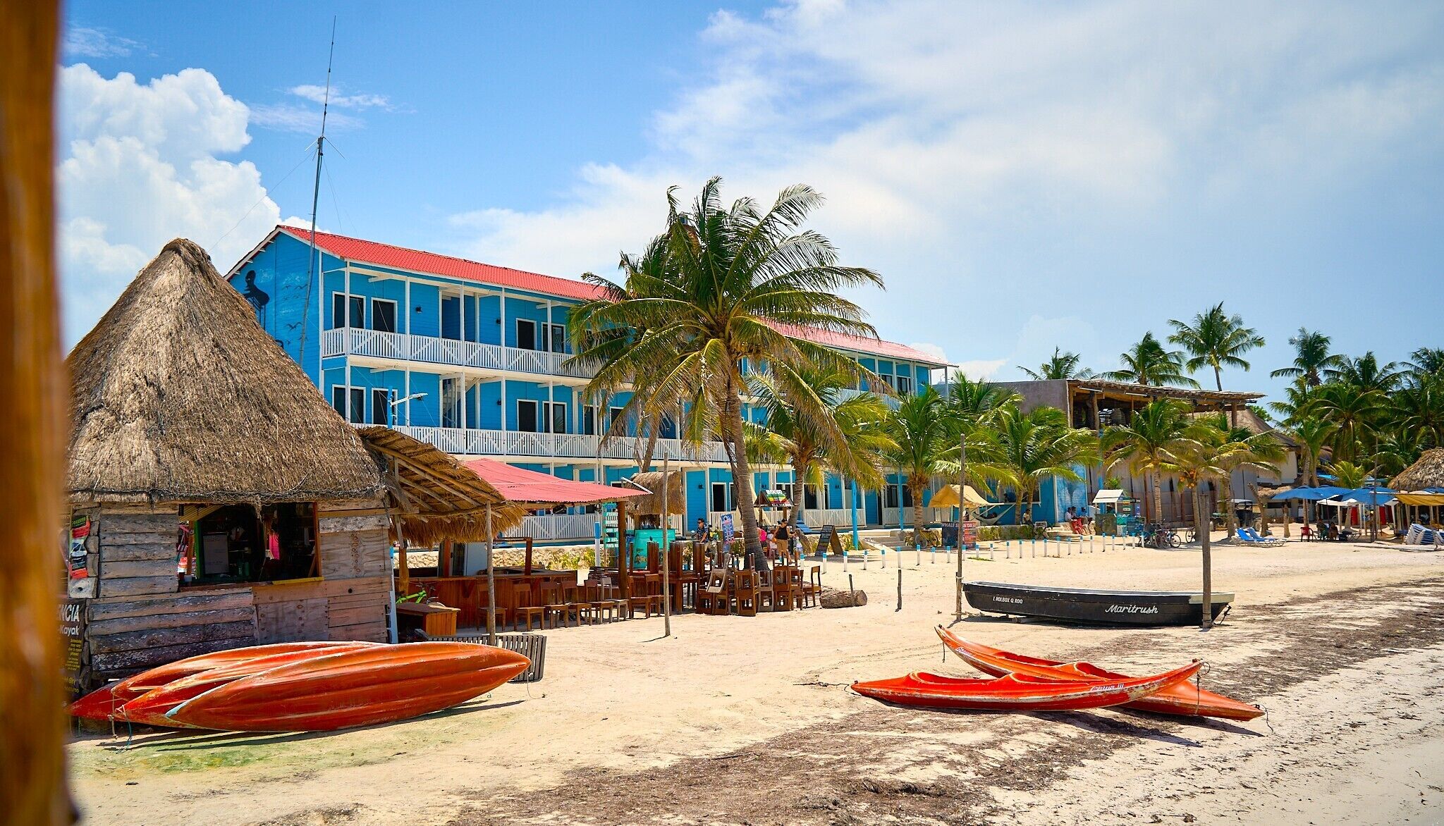 Un edificio frente a la playa con techo de paja, un edificio azul con balcones y varios kayaks rojos sobre la arena.