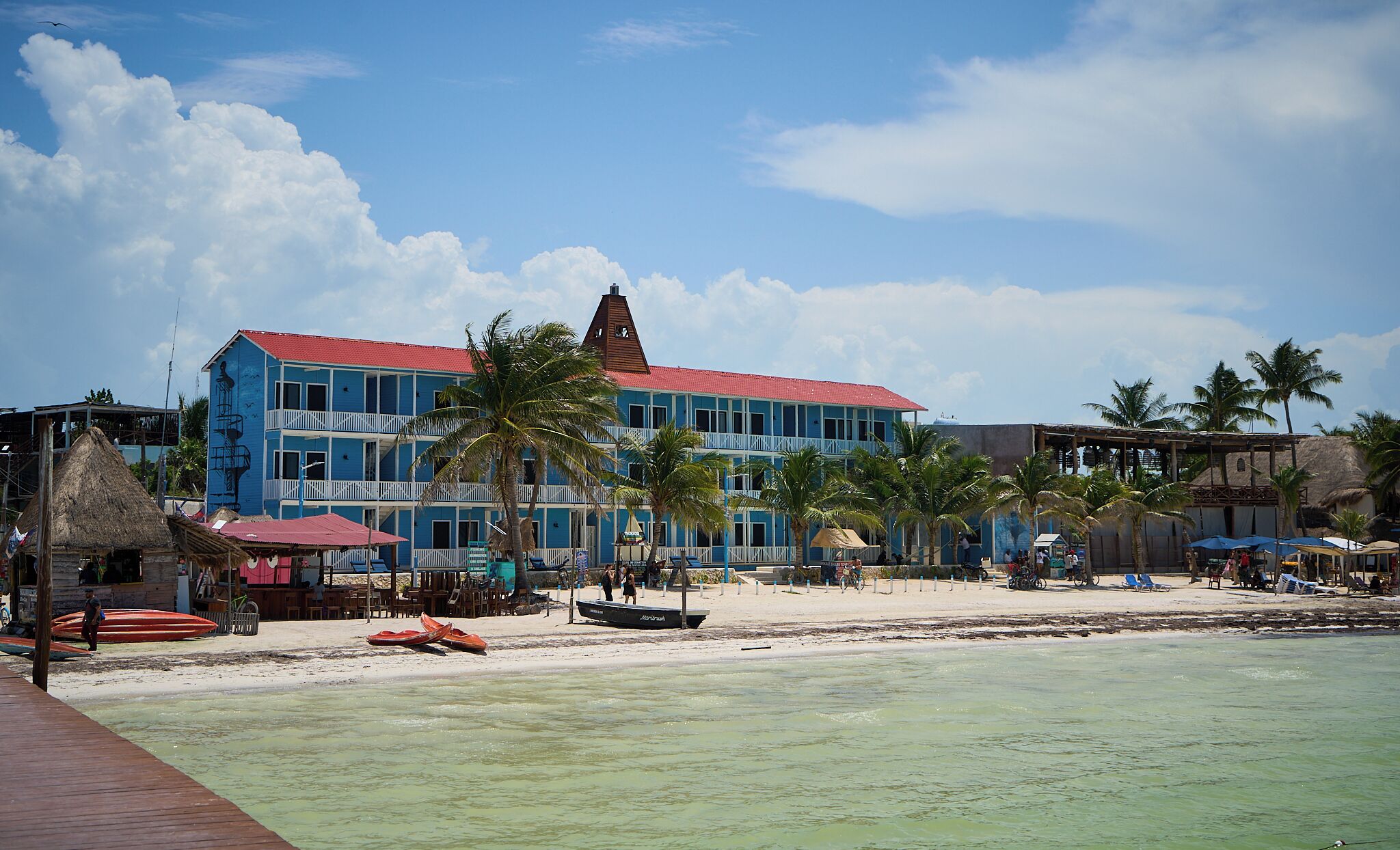 Un hotel frente al mar con varios balcones, techo rojo y palmeras.