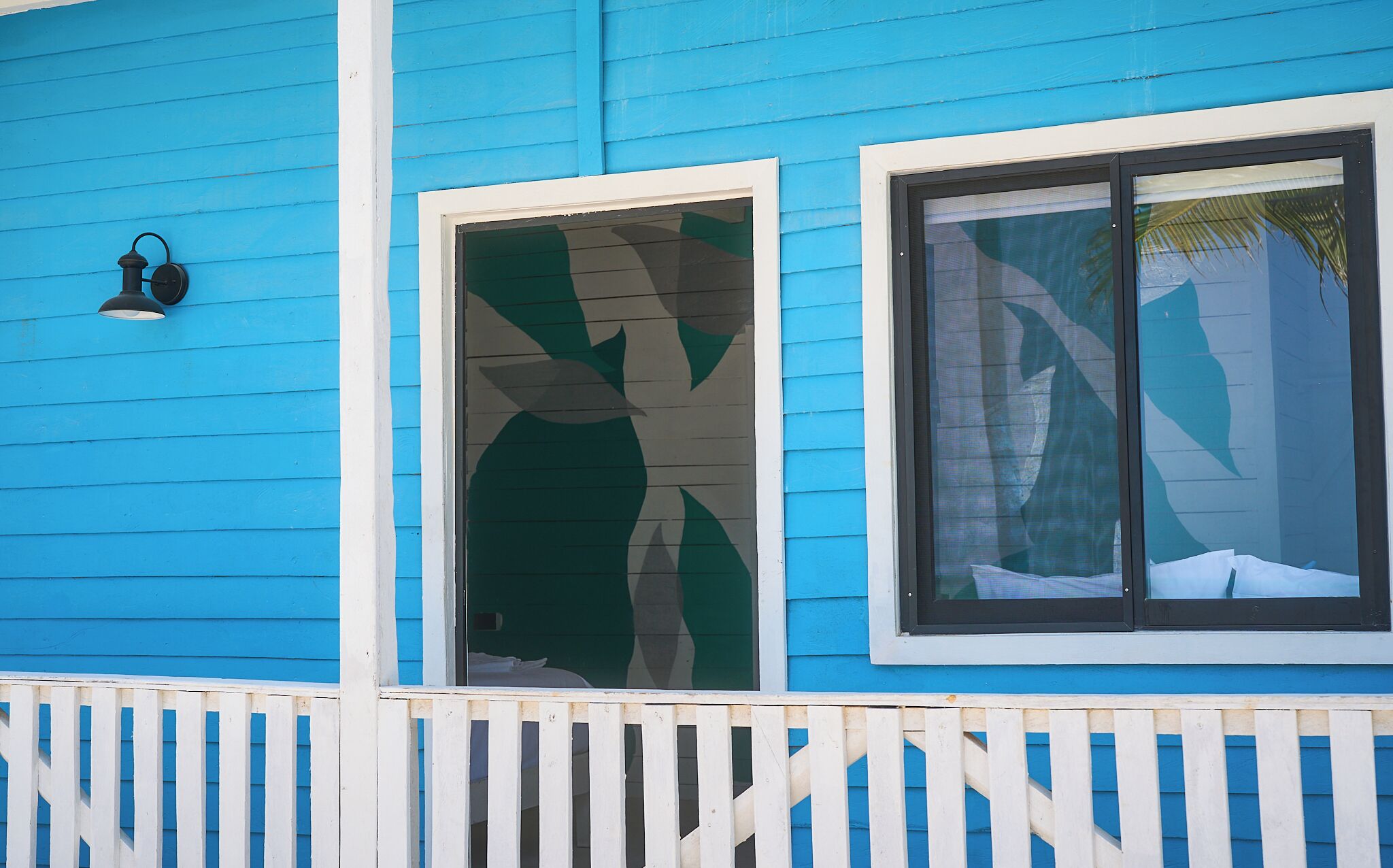 Ein blaues Haus mit weißem Zaun, ein Fenster mit grünem Blattmuster und eine schwarze Wandleuchte.