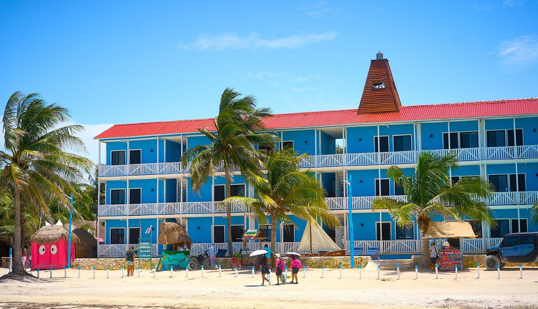 A multi-story hotel with balconies, a red roof, and palm trees in front.