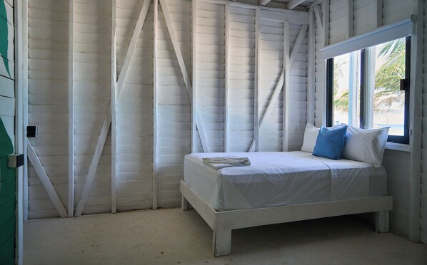 A single bed with white bedding and two pillows, a window with a view of palm trees, and white wooden walls.