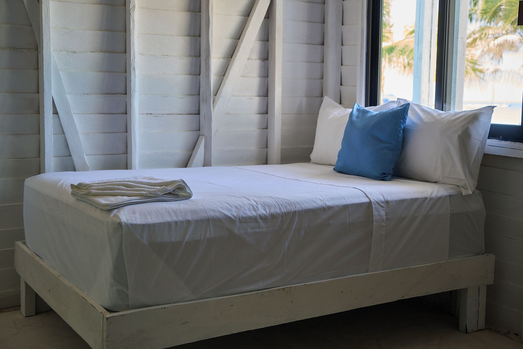 A white wooden bed with a mattress and pillows, a window with a view of palm trees, and a white wall.