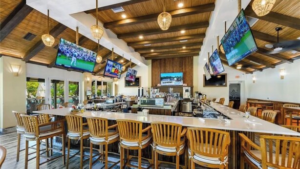 Uma área de bar com banquetas de madeira, um balcão comprido e várias televisões de tela plana instaladas no teto.