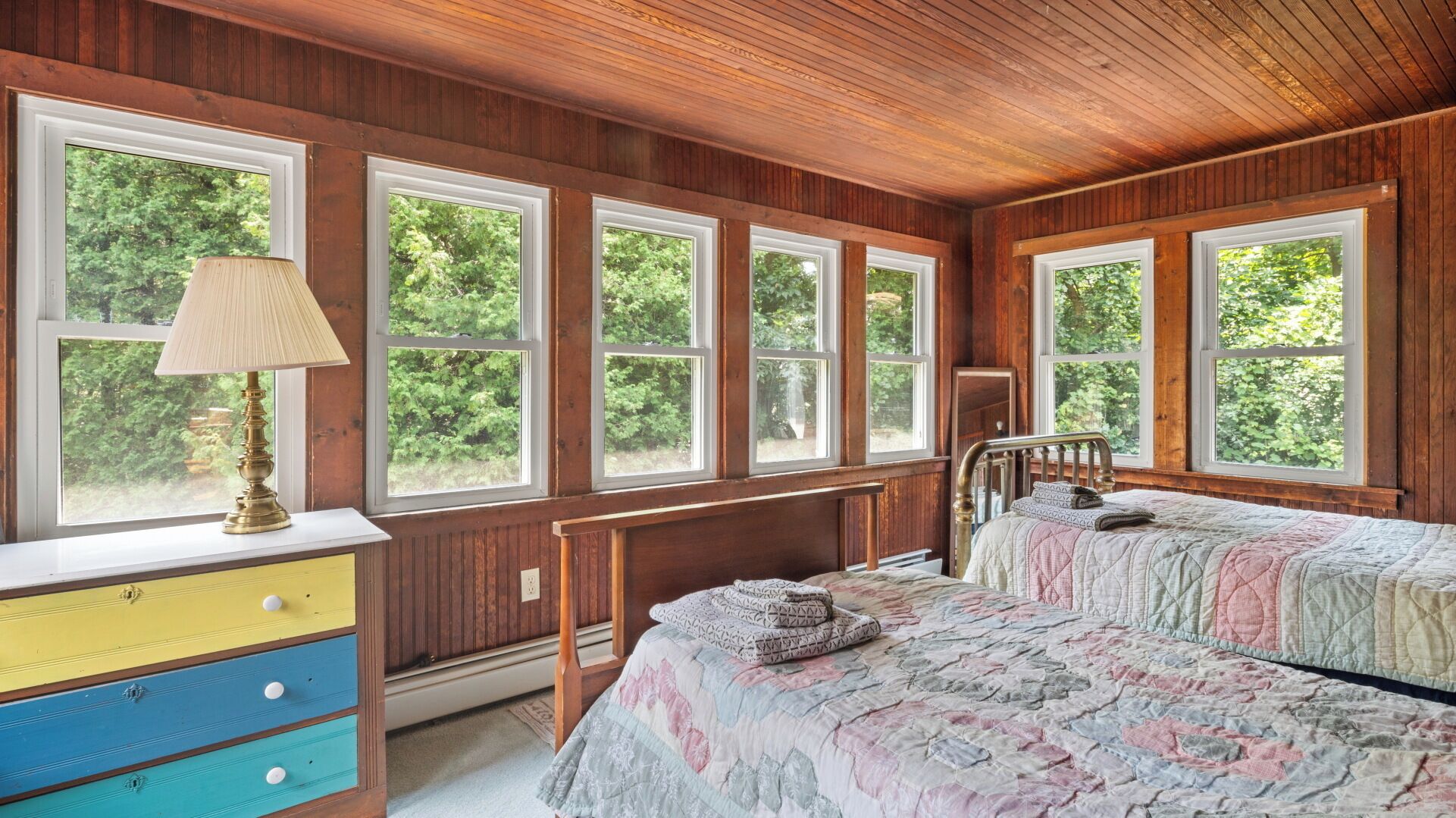 A bedroom with a wooden ceiling, large windows, a bed with a quilt, a bedside table, and a lamp.