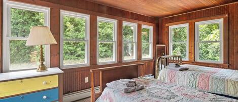 A bedroom with a wooden ceiling, large windows, a bed with a quilt, a bedside table, and a lamp.