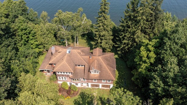 Een groot, meerlaags huis met een symmetrisch dak, omringd door dicht bos en een waterpartij.