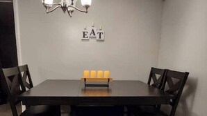 Une salle à manger avec une table en bois foncé, quatre chaises, un banc, un lustre et une enseigne fixée au mur sur laquelle on peut lire « MANGER ».