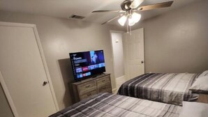 Une chambre avec deux lits, un ventilateur de plafond, un téléviseur posé sur une commode en bois et une porte.