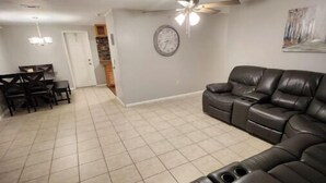 Un salon avec un canapé d’angle, un coin repas avec une table et des chaises, un ventilateur de plafond et une horloge au mur.