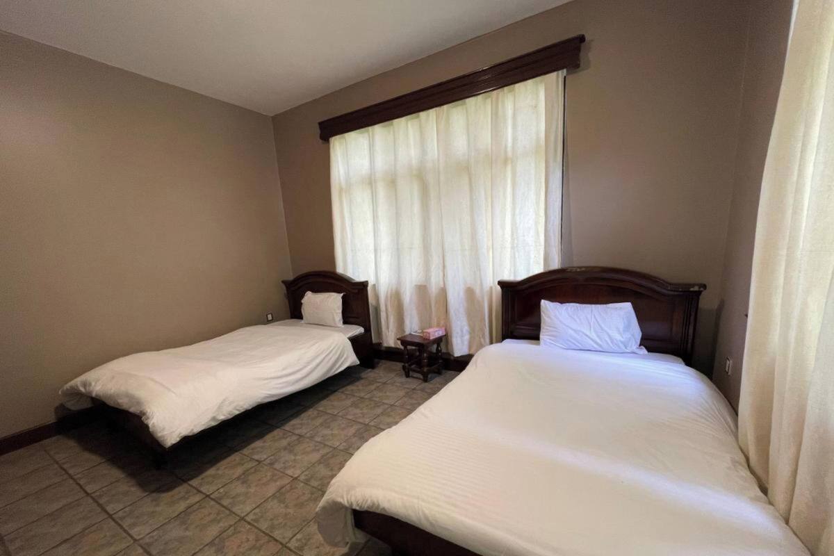 Quarto de hotel com duas camas de solteiro, uma mesinha e janela com cortinas.