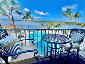 Ein Balkon mit zwei Stühlen und einem kleinen runden Tisch, mit Blick auf einen Swimmingpool und ein Gewässer mit Palmen.