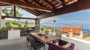 An outdoor patio with a wooden table, chairs, and a grill. There is a stone wall and a view of the sea. - Villa Karla in Poljane (Opatija)