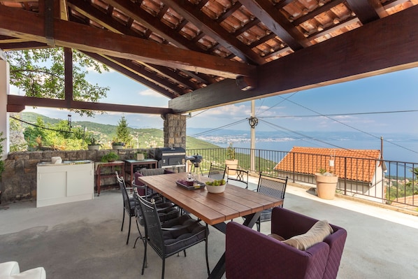 Patio esterno con tavolo e sedie in legno, e un barbecue. Si nota un muro in pietra e vista sul mare.