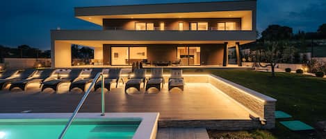 A modern two-story house with a swimming pool, outdoor seating, and a well-lit patio area.