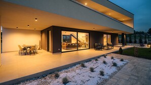Een modern huis met een overdekt terras, buitenmeubels en een zichtbare trap die naar binnen leidt.