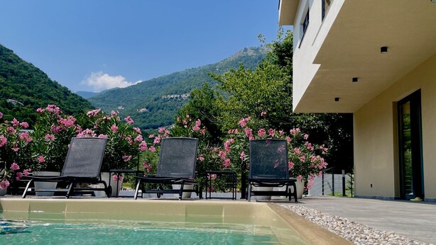 Ein Pool mit klarem Wasser, umgeben von Liegestühlen und rosa Blumen, mit Blick auf eine Berglandschaft im Hintergrund.
