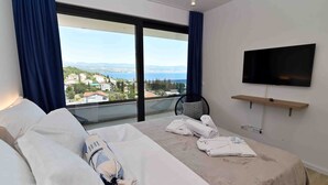 Ein Hotelzimmer mit Bett, Wandfernseher und Blick auf Stadt und Meer durch große Fenster.