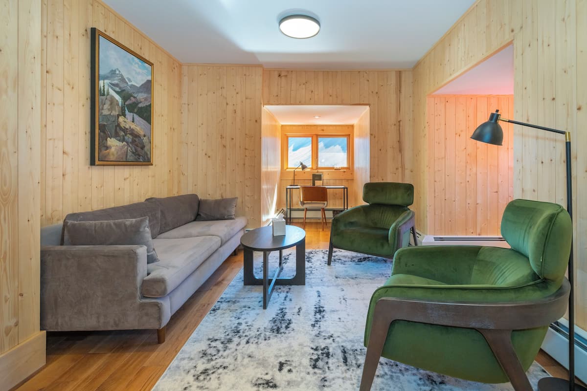 Una habitación acogedora con paredes de madera, un sillón verde, un sofá gris y una pequeña mesa con una lámpara.