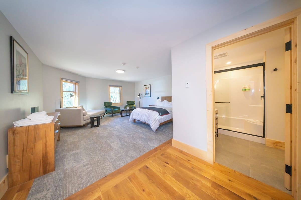 Un dormitorio moderno con cama, escritorio, silla y una mesita. Hay una puerta que da a un baño.