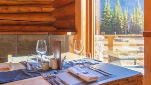 Interior de una cabaña de madera con una mesa de comedor para dos personas, mostrando una vista de un paisaje montañoso a través de un gran ventanal.