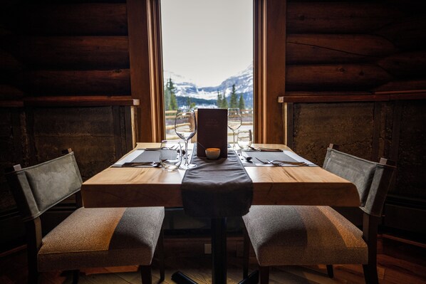 A wooden dining table with two chairs, a menu stand, and a view of snow-capped mountains through a window.