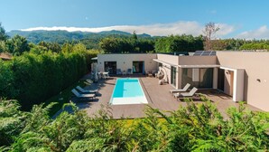 Un área de piscina moderna al aire libre con sillones de descanso, una zona de estar cubierta y vistas a montañas al fondo.