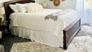 Una cama bien hecha con una colcha blanca y varias almohadas, un cabecero de madera oscura y una pared decorada con varios sombreros.