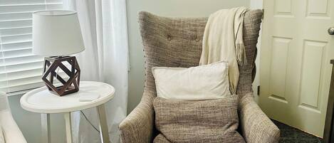 Una habitación con un sillón color beige, una mesita redonda con una lámpara, un cuadro en la pared y una puerta.
