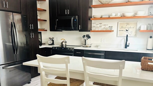 Una cocina moderna con armarios oscuros, electrodomésticos de acero inoxidable y una zona de comedor blanca con sillas de mimbre.