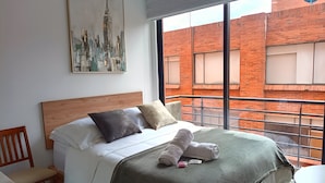 A hotel room with a large bed, a wooden headboard, a cityscape painting, and a view of a brick building through a large window. - Bright Studio with Balcony 615 (Bogotá)