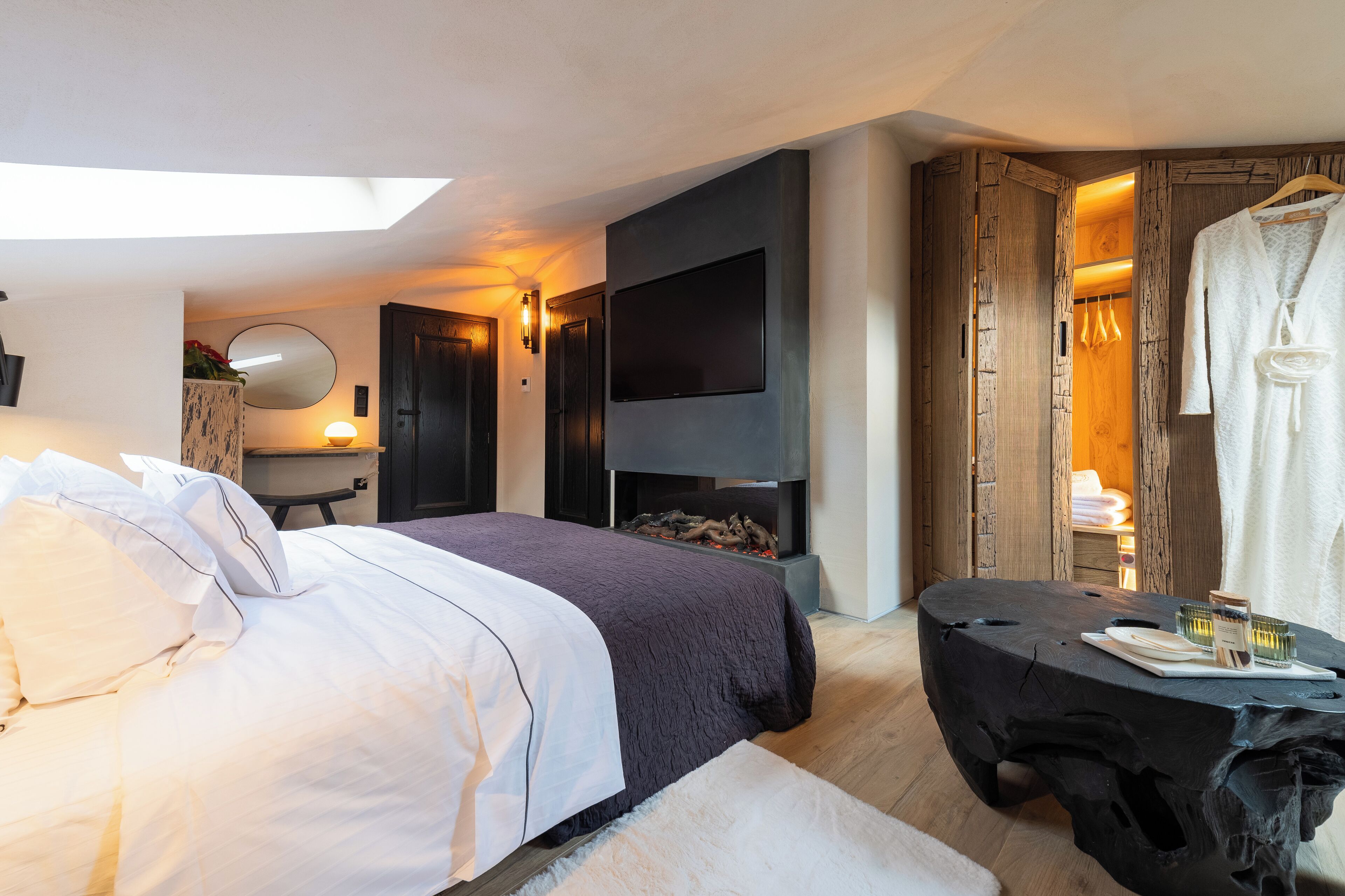 A modern hotel room with a large bed, a flat-screen TV, a fireplace, and a wooden wardrobe.