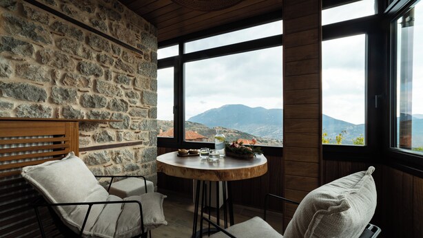Ein gemütliches Zimmer mit Steinwänden, einem runden Holztisch mit Bergblick, zwei Stühlen mit Kissen und einer hölzernen Bank.